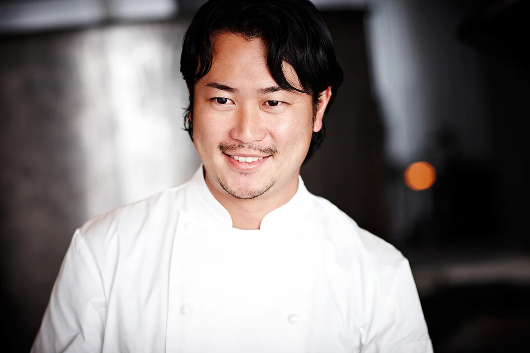 A man wearing a white chef's coat smiling at the camera in an indoor setting with blurred background.