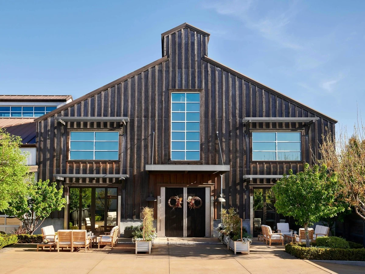 Exterior of a converted barn-style winery building with weathered vertical wood siding, modern blue-paned windows, central cupola, and outdoor tasting
