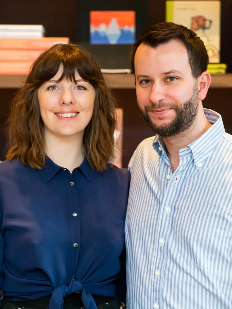 A man and a woman posing together in front of a bookshelf filled with books, consistent with the co-owners of Lucian Books and Wine.