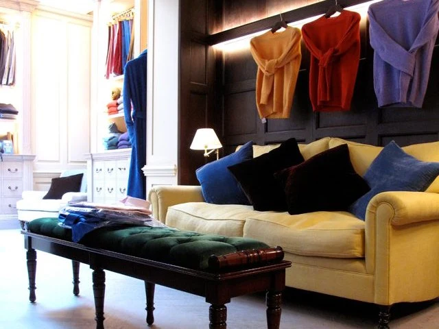 Interior of Anderson & Sheppard Haberdashery on Clifford Street, featuring a yellow sofa, green bench, and sweaters on a dark wood wall.