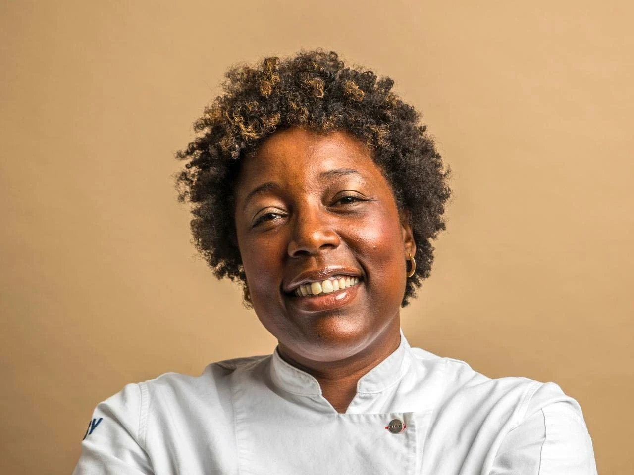 A smiling Black woman with short curly hair wearing a white chef's coat, photographed against a beige background.