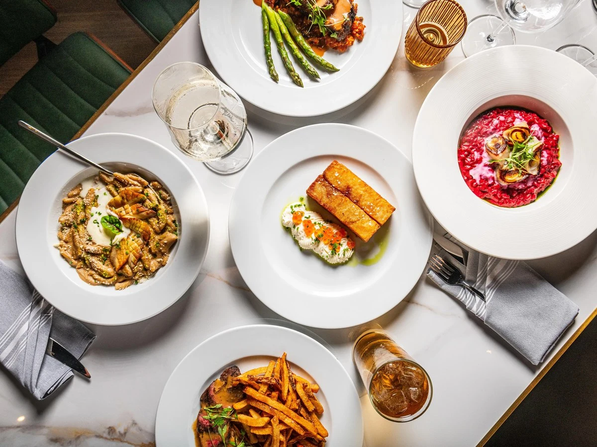 An overhead shot of a restaurant table set with multiple plated dishes including pasta, beet risotto, fried fish with roe, steak frites, and a meat.