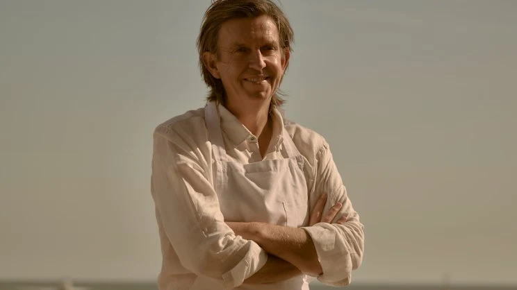 Portrait of a person in chef's whites with arms crossed against a neutral background