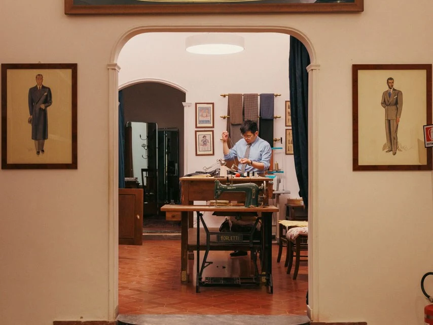 A tailor works at a Borletti sewing machine inside the Liverano & Liverano atelier, with a prominent 'LIVERANO' sign above the arched entrance.
