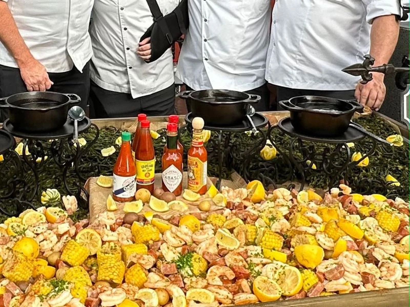 Multiple chefs in white chef coats stand behind a table displaying a seafood boil with corn, potatoes, sausage, lemon slices, and various hot sauce bottles in front.