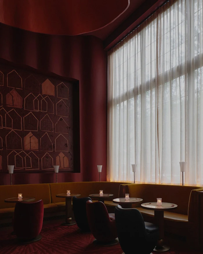 Upscale restaurant dining room with burgundy velvet banquette seating, round tables with small candles, sheer white curtains, and a decorative wall pa