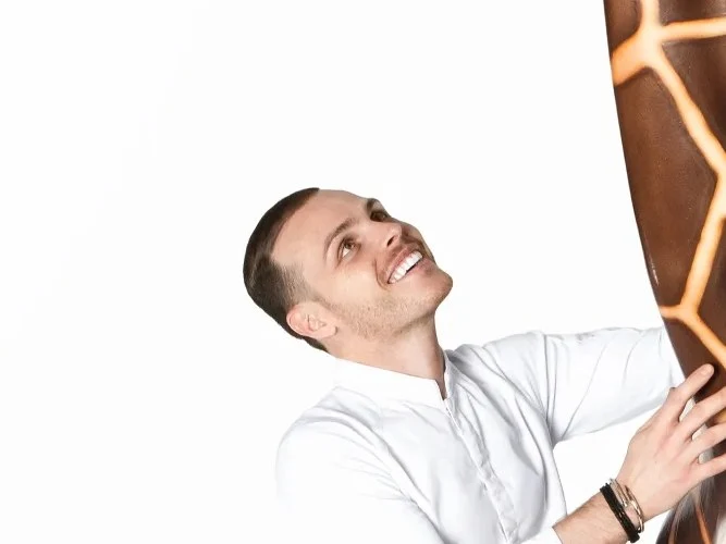 A man in a white shirt and black pants stands next to a near life-size chocolate giraffe sculpture on a white pedestal.