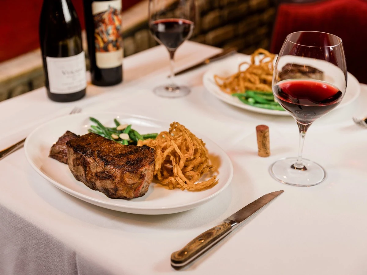 A white-tablecloth table set with a plated steak, green beans, and onion strings, alongside two glasses of red wine and two wine bottles.