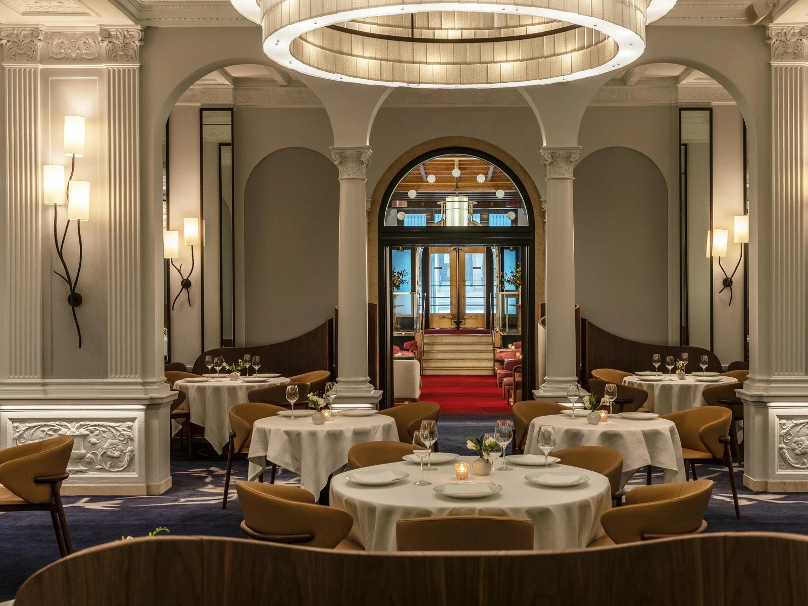 Elegant fine dining restaurant with round tables covered in white tablecloths, mustard-gold upholstered chairs, ornate Corinthian columns supporting