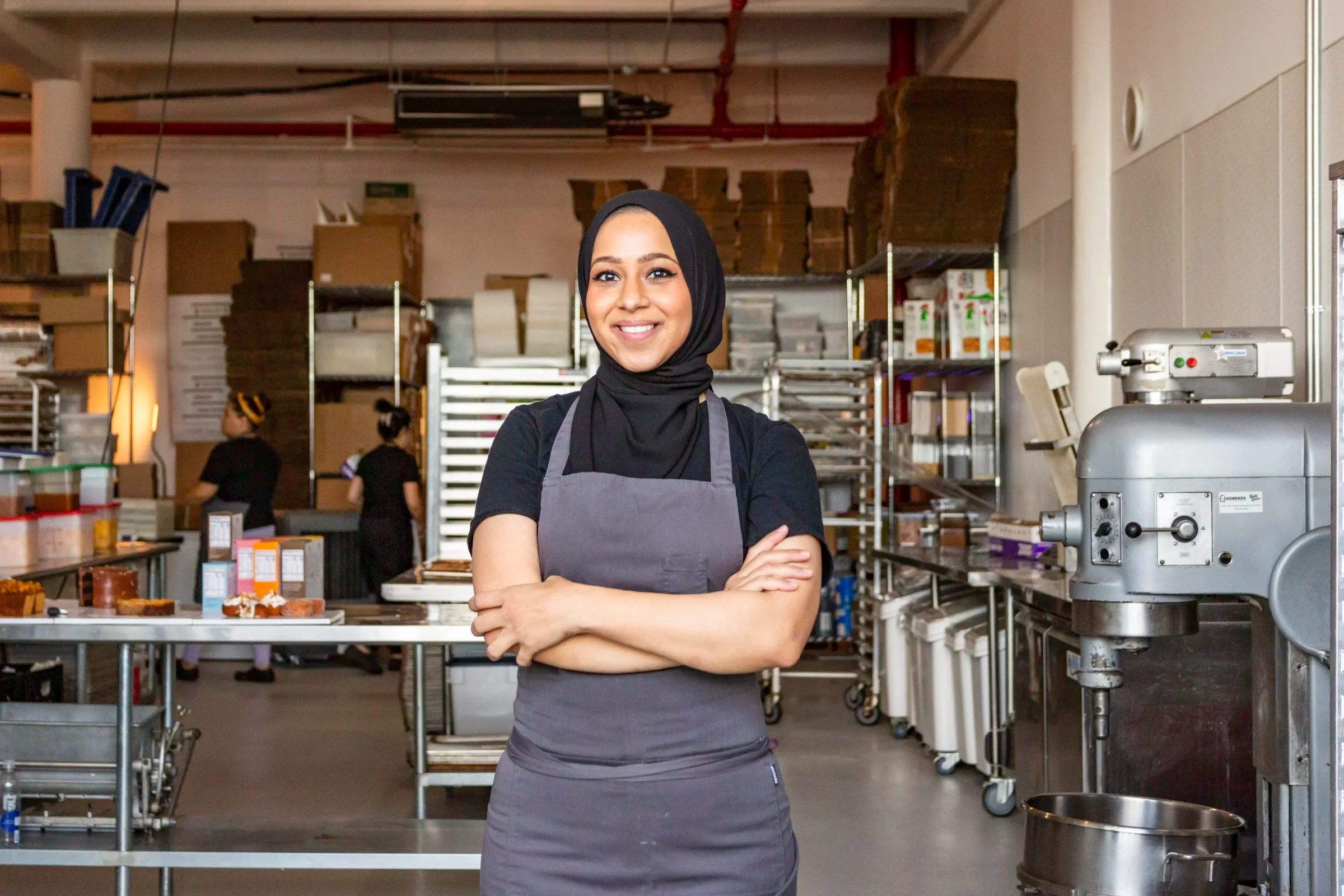 A smiling woman in a black hijab and gray apron stands with her arms crossed in a professional kitchen.