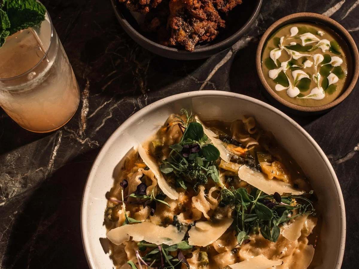 A dark marble table holds a pasta dish with cheese and herbs, a bowl of fried food with lemon and cilantro, a green soup, and a cocktail.