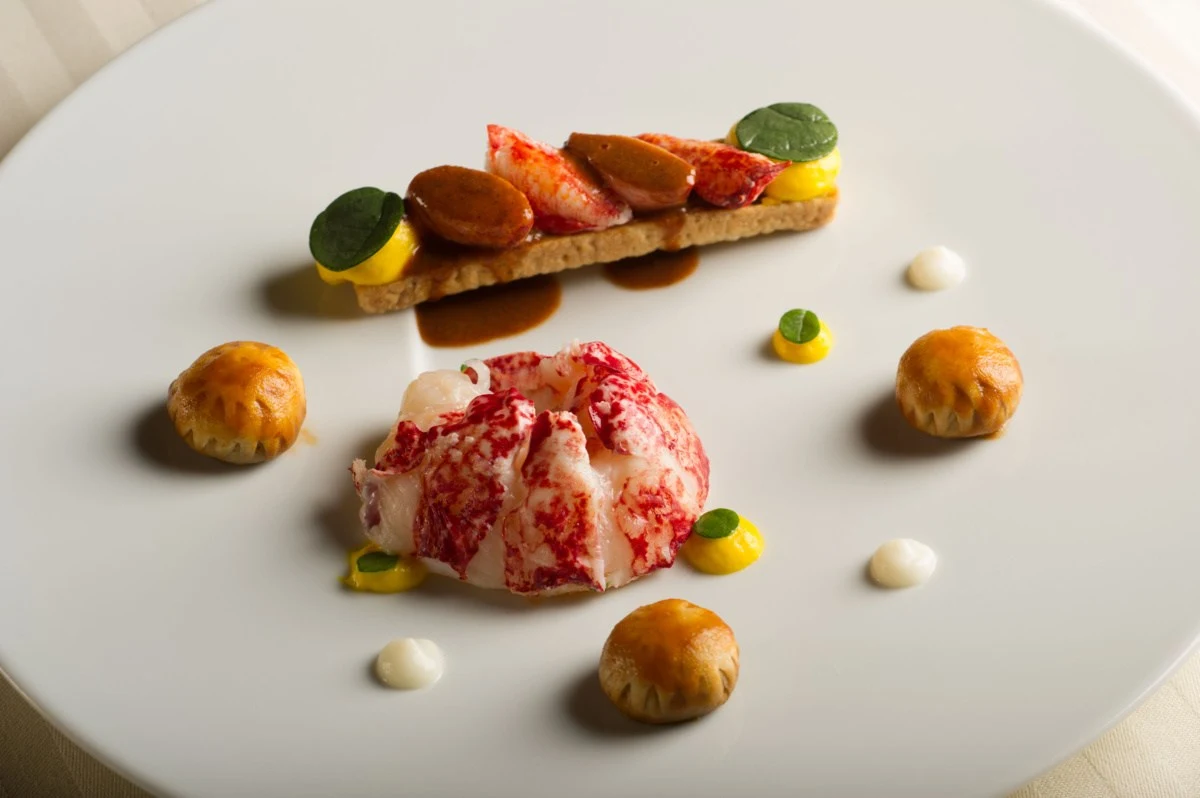 An elegantly plated fine-dining dish featuring lobster meat, crispy pastries, and colorful sauces on a white plate
