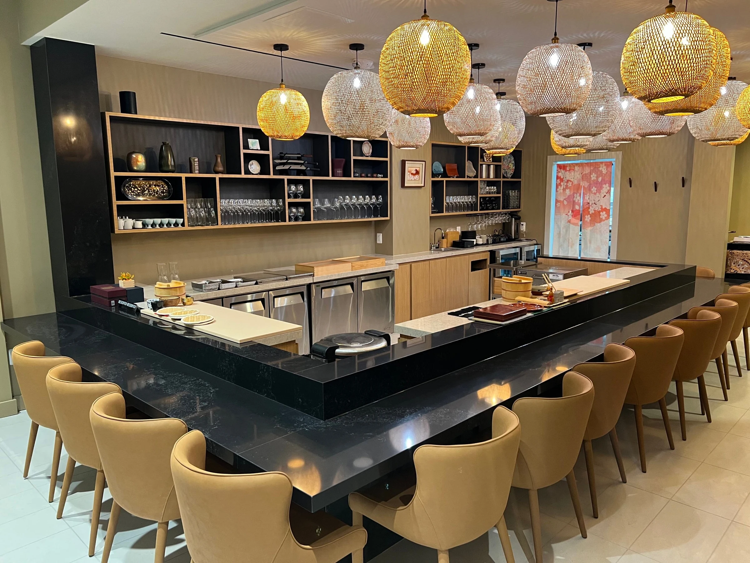 A modern sushi omakase counter with a black stone surface, tan upholstered chairs, and decorative woven pendant lights.