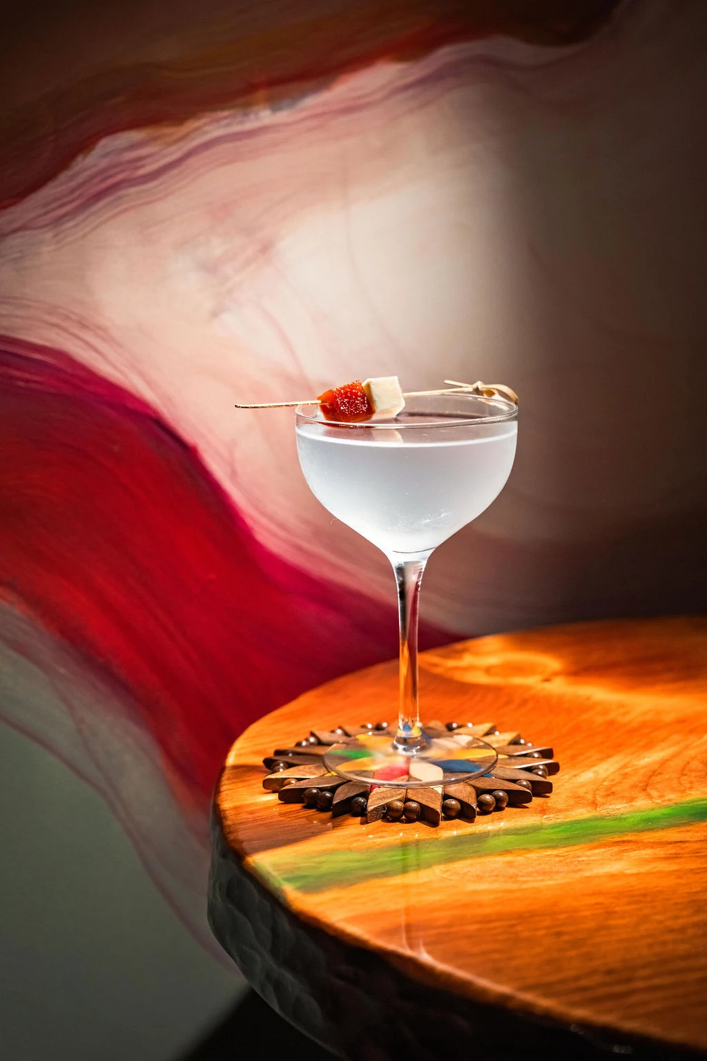 A professionally photographed cocktail in a coupe glass with a strawberry-and-marshmallow garnish, set on a decorative wooden coaster against a colorf