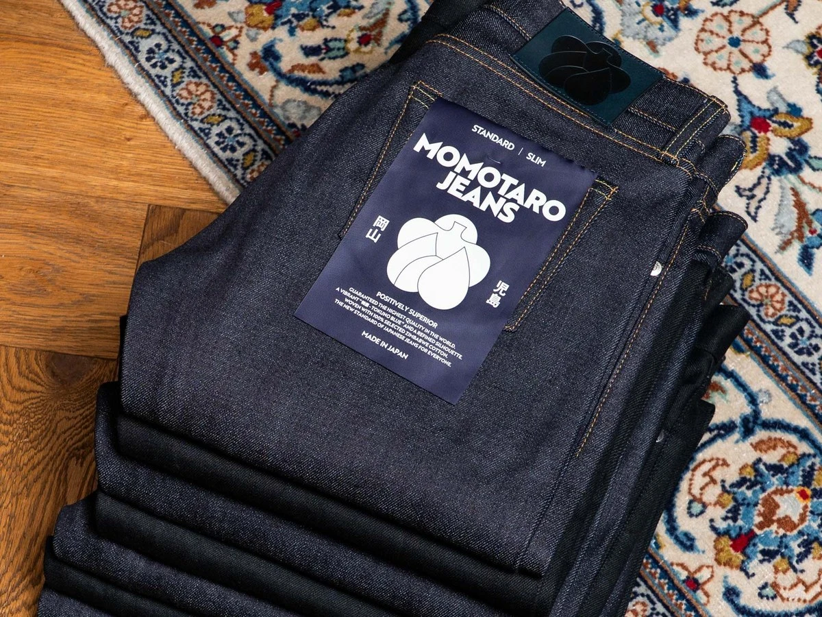 A stack of folded dark indigo selvedge denim Momotaro Jeans with a "STANDARD | SLIM" hang tag, featuring the brand's peach logo and "MADE IN JAPAN"