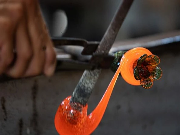 A close-up shows a glassblower's hand holding metal jacks, shaping molten orange glass into a flower-like form on a blowpipe.