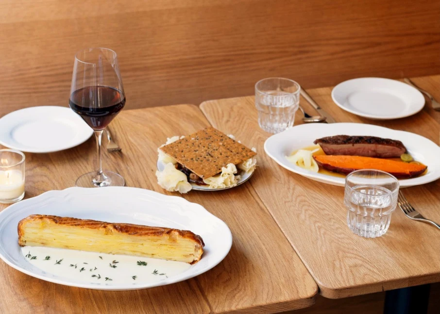 Editorial dining table scene showing plated dishes including what appears to be a layered potato/sunchoke tart, sausage with vegetables, and a s'more 