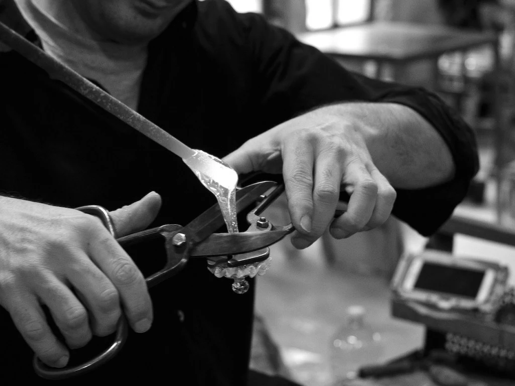 Close-up black-and-white photo of a craftsman's hands using large metal shears to shape molten glass on a blowpipe.