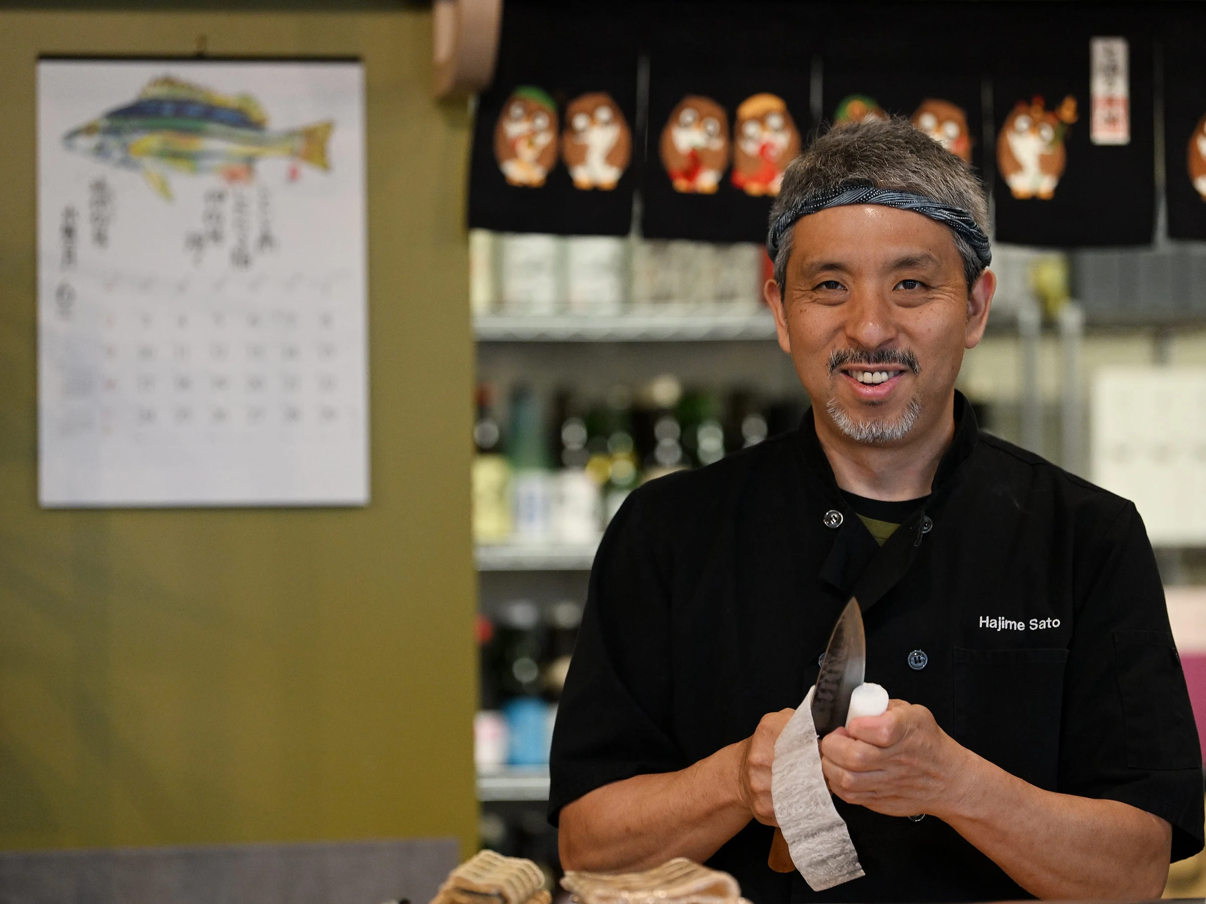 A man wearing a black chef's coat with the name Hajime Sato embroidered on it stands smiling in a restaurant, holding a knife wrapped in cloth, with Japanese artwork and masks displayed behind him.