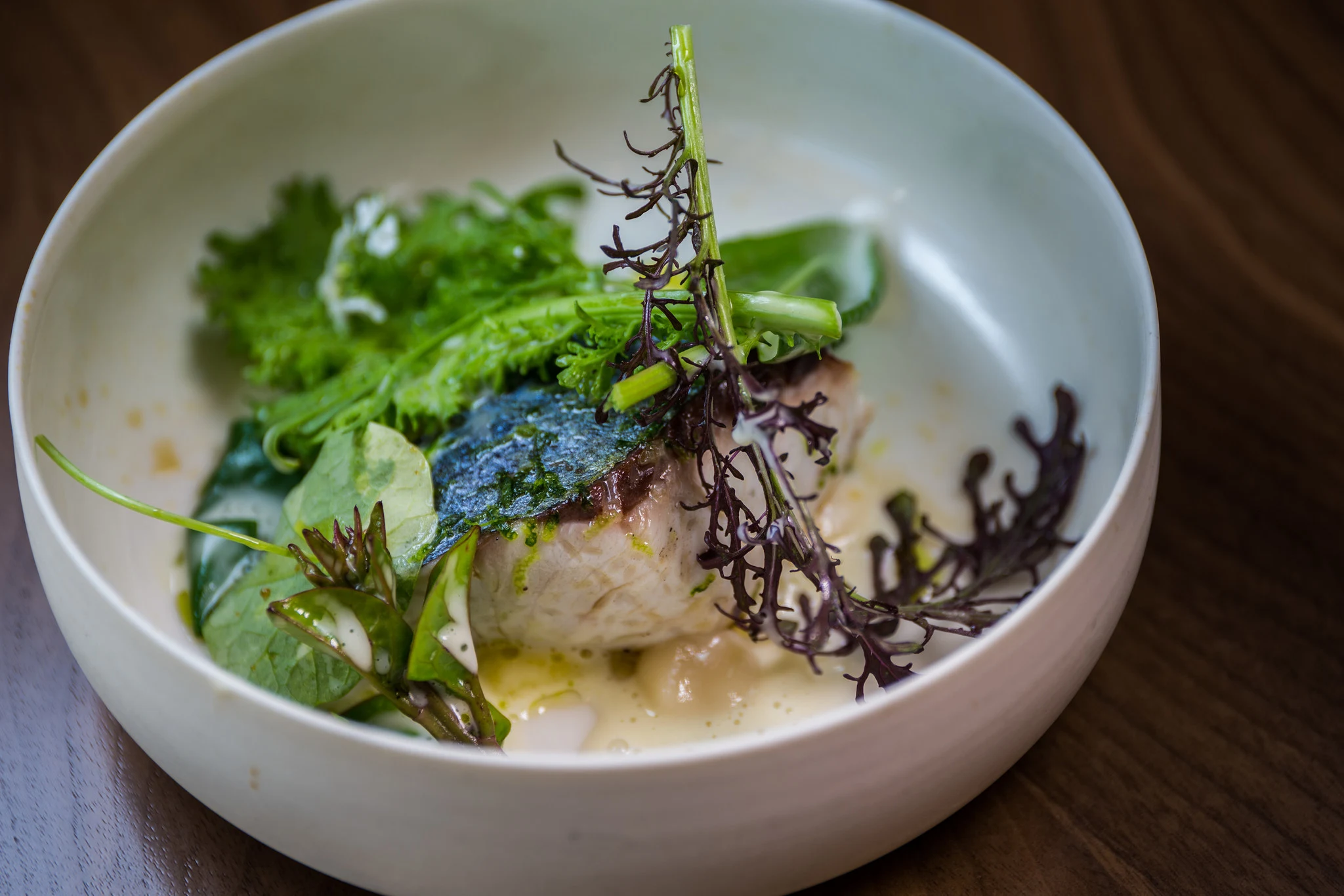 A plated seafood dish featuring Japanese tai (sea bream) with crispy skin, garnished with seaweed, fresh greens, and herbs in a white bowl, set in a c