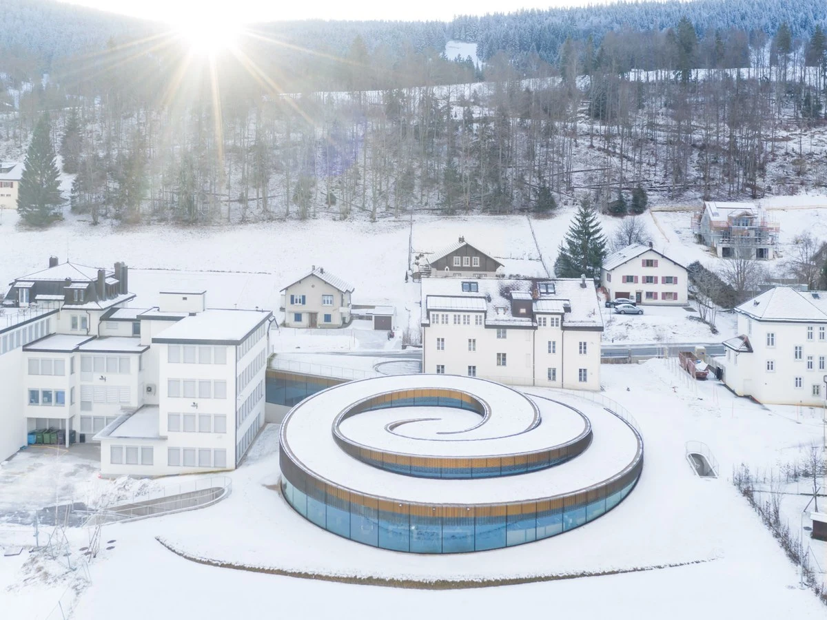 Aerial drone photograph of the Musée Atelier Audemars Piguet in winter snow, showing the BIG-designed circular glass-and-wood spiral building set