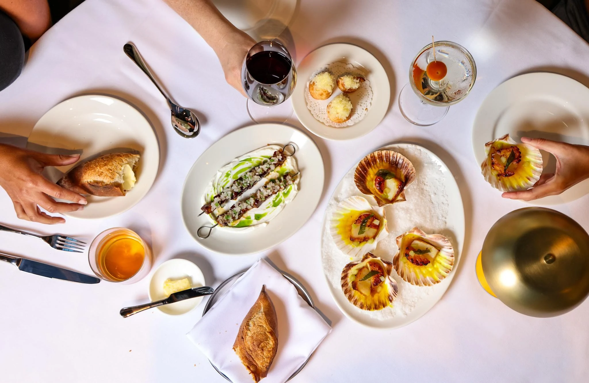 Overhead shot of multiple starter dishes on a white marble table, including scallops in shells, gougères, and other small plates with wine glasses; so