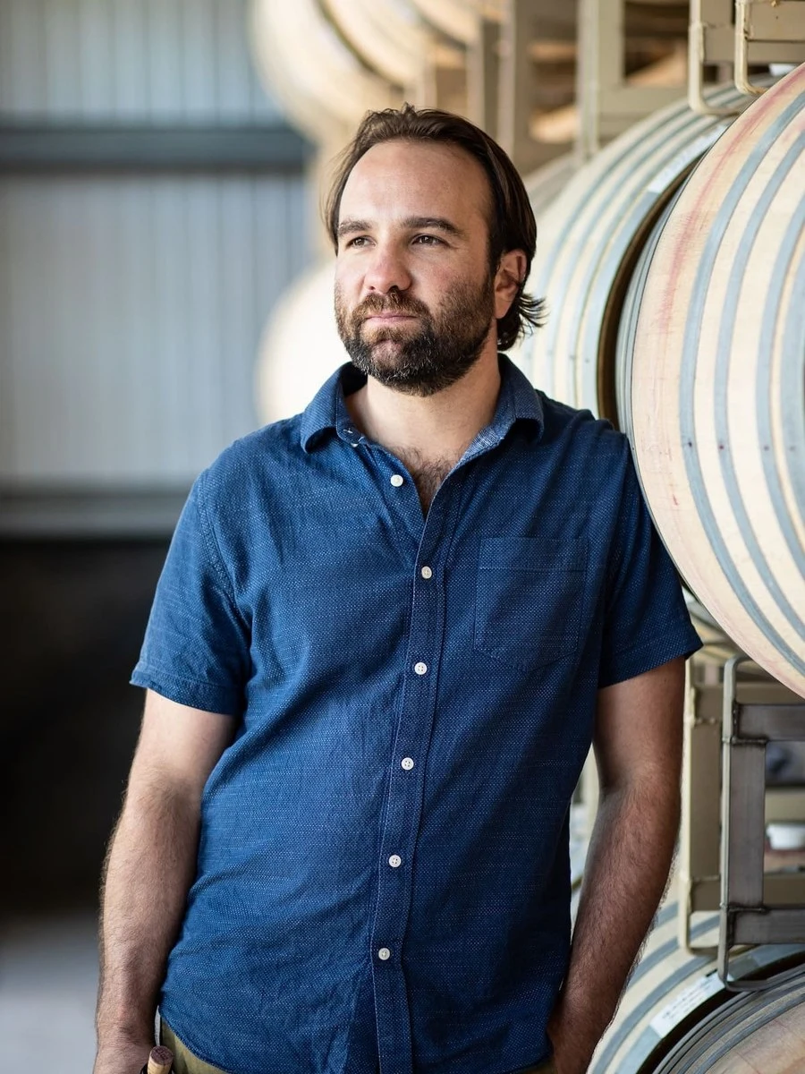 A bearded man wearing a dark blue denim shirt poses next to striped wooden wine barrels in an industrial winery setting.