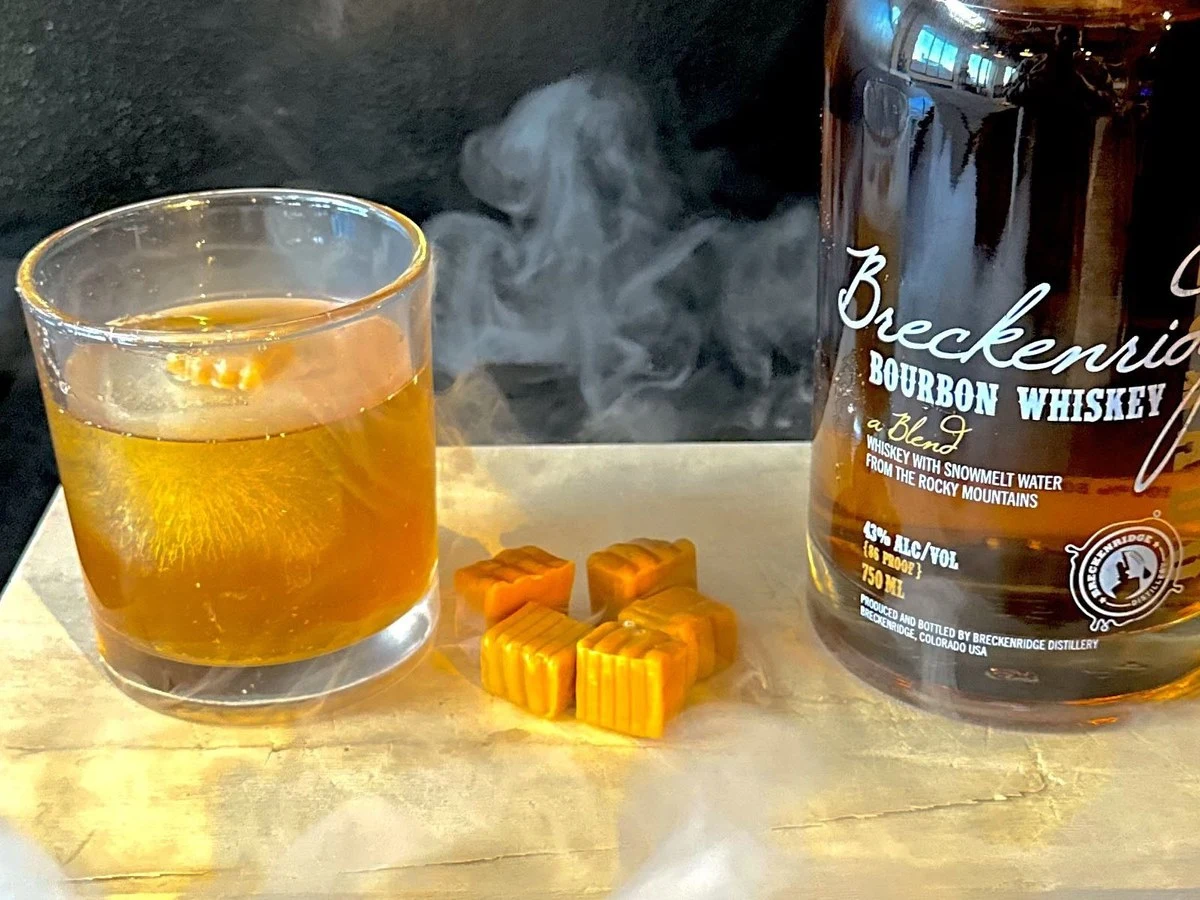 A glass of amber liquid with ice, a bottle of Breckenridge Bourbon Whiskey, and five orange candies on a light surface with smoke.