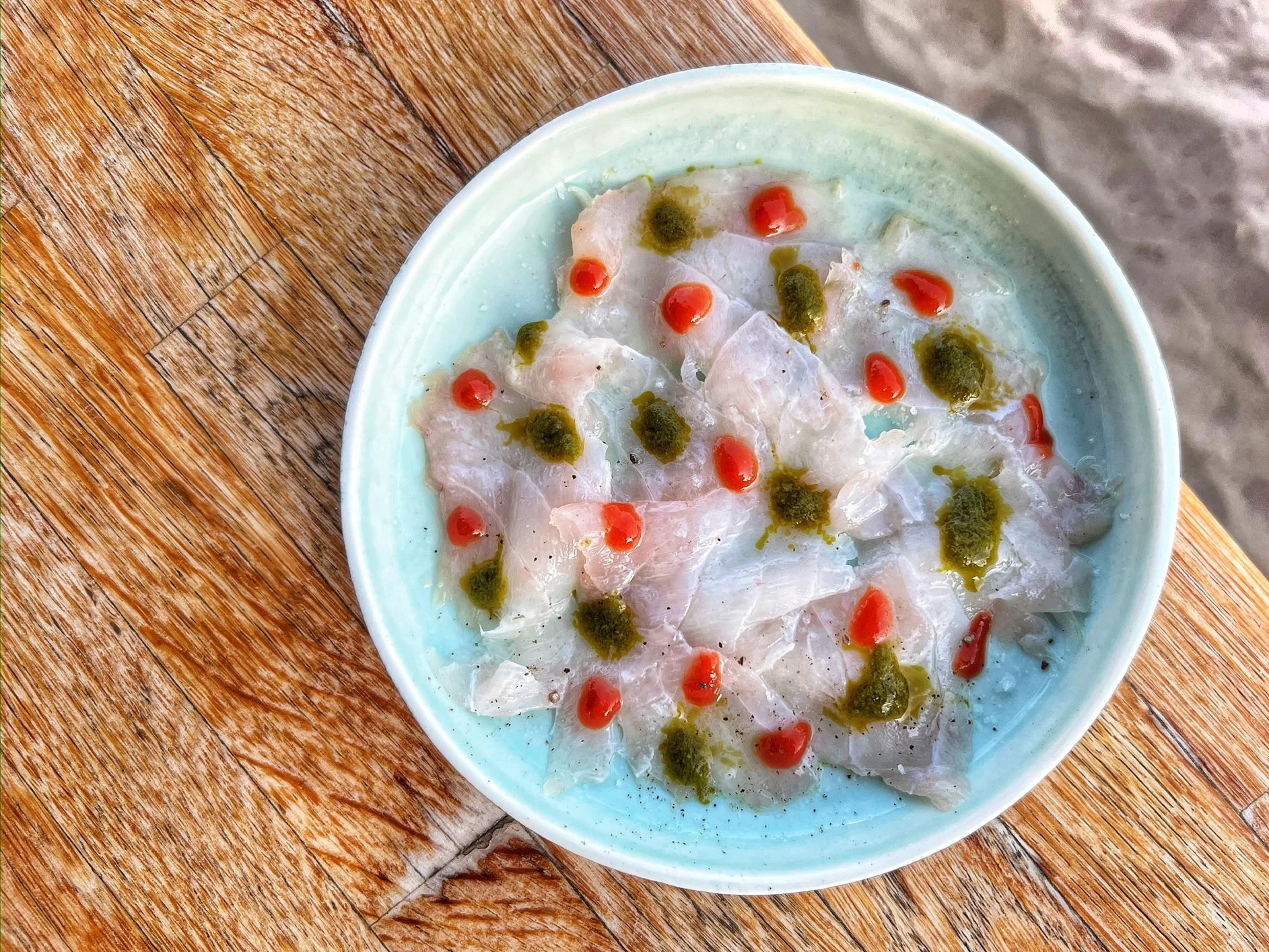 A light blue ceramic bowl containing thin slices of raw fish (likely a crudo or sashimi preparation) garnished with dots of red sauce and green herb o