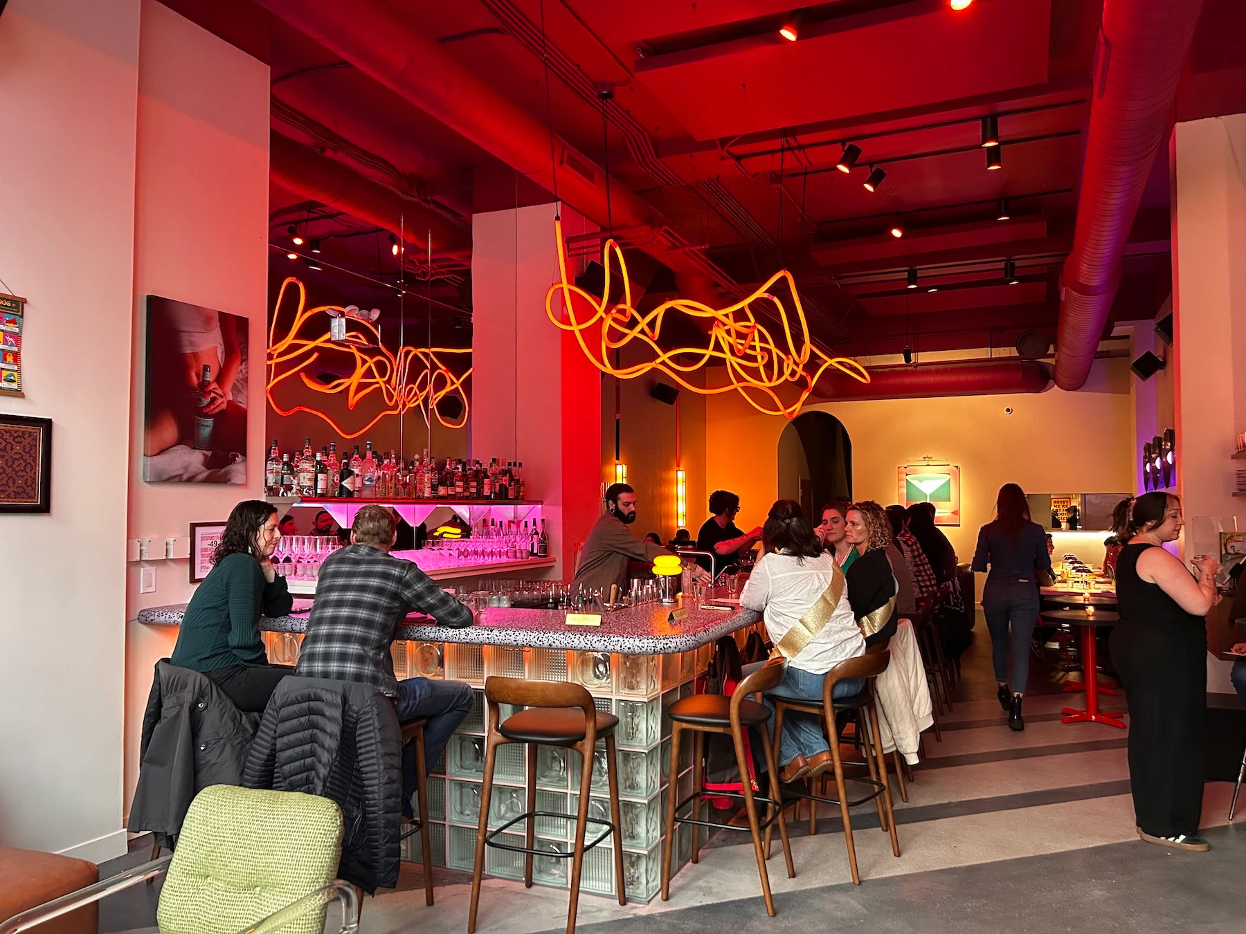 A modern cocktail bar interior with distinctive orange neon art installation overhead, glass-block bar construction, pink-lit back bar, and patrons se