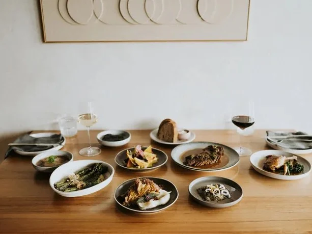 A wooden table is laden with an array of elegantly plated dishes, including roasted meats, asparagus, and various salads, alongside wine glasses.