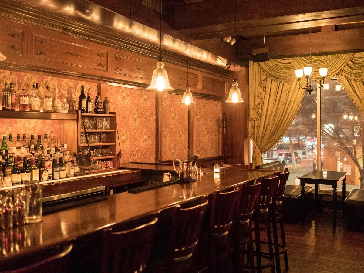 A dimly lit bar interior with a long wooden counter, bar stools, shelves of liquor bottles, and gold draped curtains next to a window.