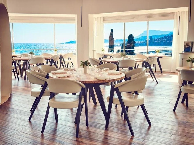 A bright, modern restaurant dining room with round wooden tables, cream upholstered chairs, and floor-to-ceiling sea-facing windows revealing a Medite.