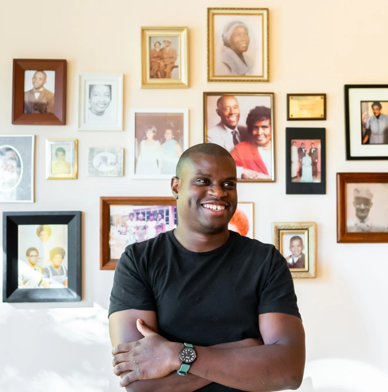 A smiling man in a black t-shirt with his arms crossed stands in front of a wall adorned with numerous framed family photographs.
