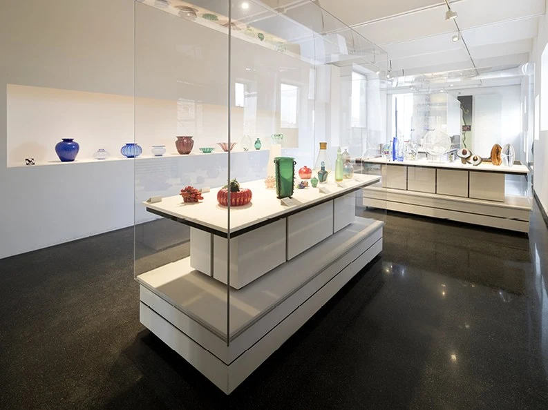 A bright, modern museum gallery interior displaying colorful Murano glass objects in glass-enclosed pedestals and wall shelves.