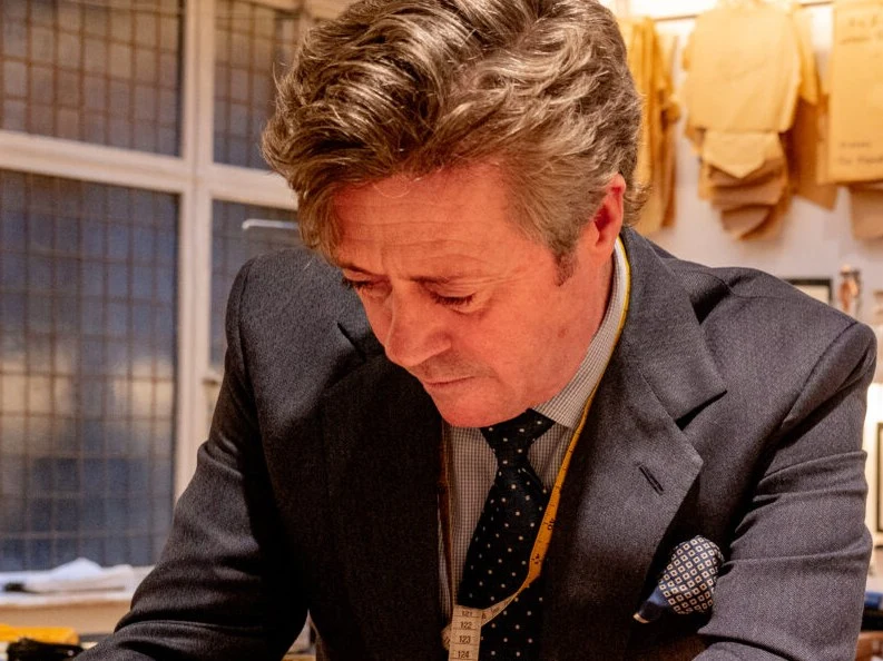 A man in a dark suit and tie leans over a work table, studying fabric samples in a tailoring workshop with windows and hanging materials visible in the background.