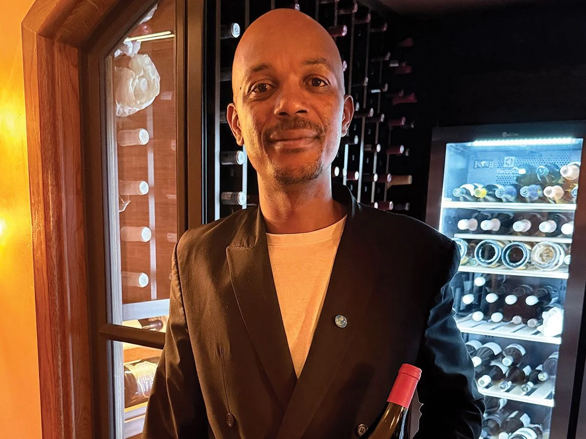 A man wearing a black double-breasted blazer over an orange shirt poses in front of glass-fronted wine refrigerators filled with bottles.