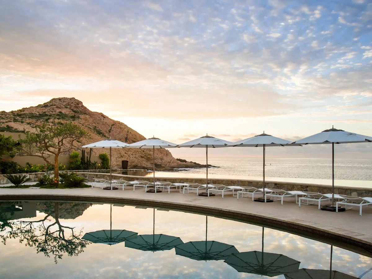 An infinity pool terrace with white sun loungers and umbrellas overlooks the Sea of Cortez against a rocky Baja hillside at golden hour.