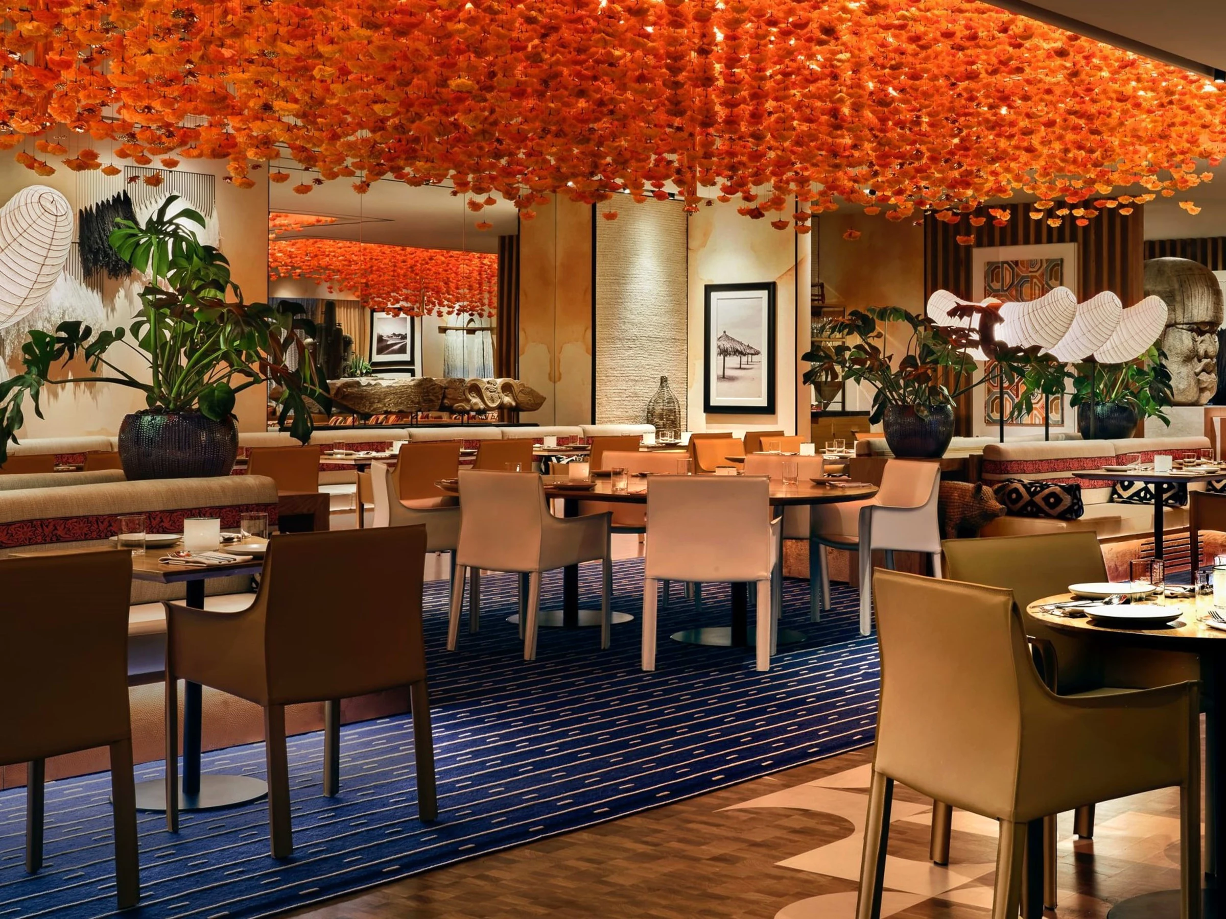 A modern restaurant dining room with tan leather chairs, tables set with place settings, a blue striped carpet, and thousands of orange paper flowers hanging from the ceiling as a dramatic art installation.