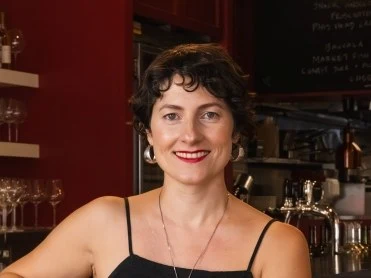 A smiling woman with curly dark hair wearing a black sleeveless top and necklace, photographed indoors at what looks like a wine bar or restaurant.