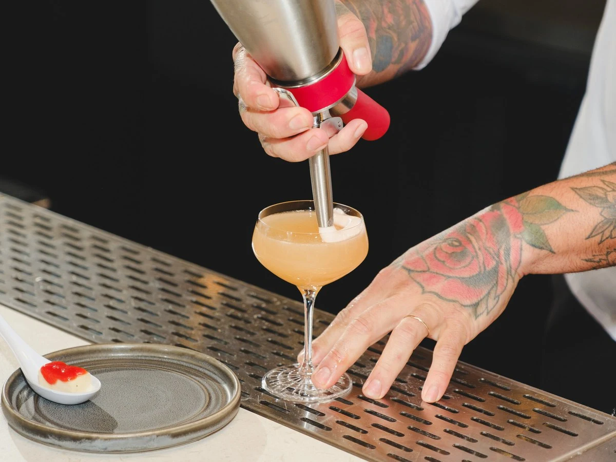 A close-up of a bartender's tattooed hands, one holding a metal cocktail dispenser pouring a creamy drink into a coupe glass, the other steadying.