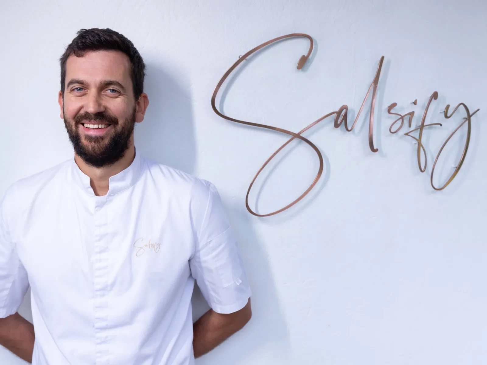 Chef in white uniform smiling beside a wall sign reading "Salsify.