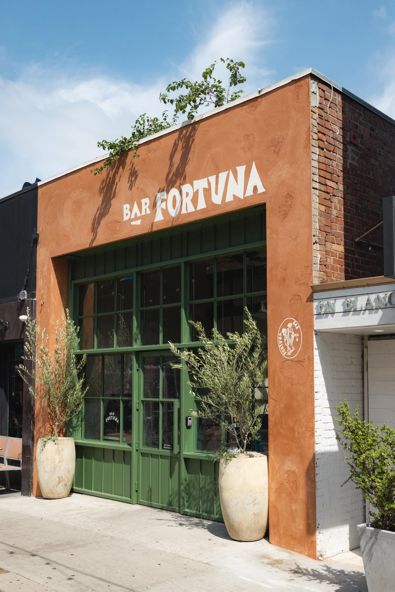 Exterior facade of Bar Fortuna in Santa Monica with prominent signage reading 'BAR FORTUNA' on an orange stucco facade with green doors and planters; 