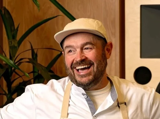Man in beige overalls and white cap smiling in front of a large speaker system in a wood-paneled room with a plant.