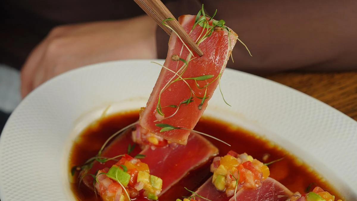 High-end seared tuna dish being lifted with chopsticks over a soy-based sauce, presented in fine-dining style