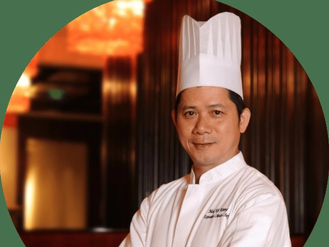 Professional portrait of an Asian male chef in white chef's uniform and toque standing in front of a wood-paneled interior with warm lighting; source 