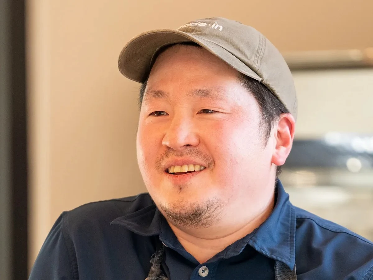 A smiling man wearing a gray baseball cap and dark denim apron stands at a counter in a professional kitchen setting.