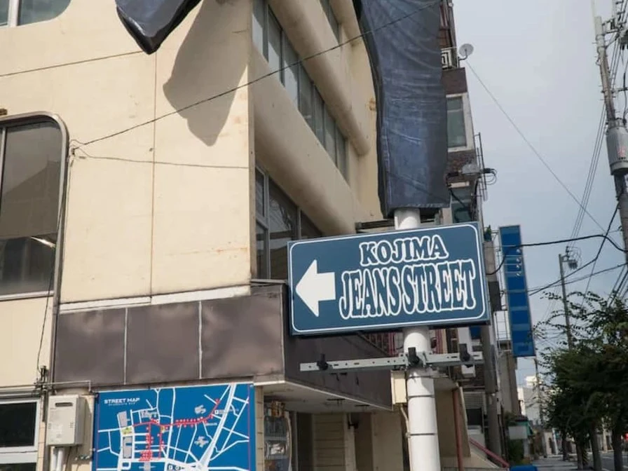 A giant denim jeans sculpture hangs from a building corner above a blue street sign pointing left, reading "KOJIMA JEANS STREET."