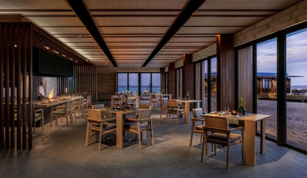 Modern upscale restaurant dining room with wooden tables and chairs, slatted ceiling, open kitchen visible through vertical slats on left, floor-to-ce
