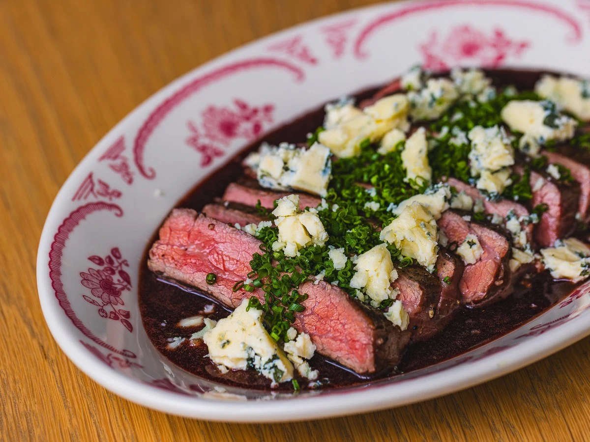 A plated dish of sliced medium-rare steak topped with crumbled blue cheese and fresh parsley on a decorative plate with pink floral pattern; the sourc